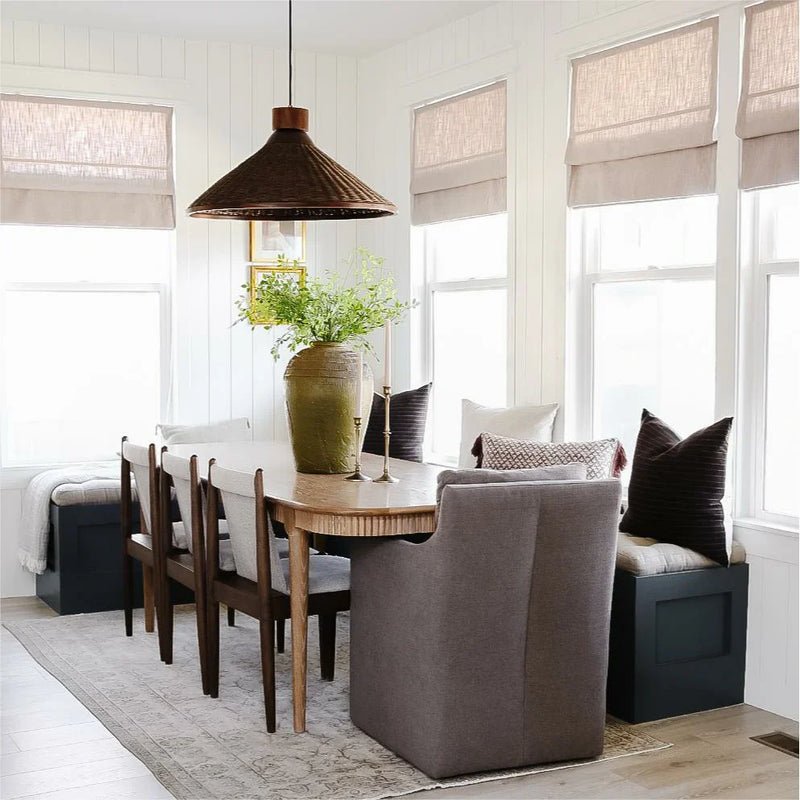 A cozy modern farmhouse dining room with white shiplap walls, centered around an oval wooden table under a dark brown woven pendant light, adorned with a tall distressed green vase of greenery and flanked by upholstered seating and multiple windows dressed in light linen Roman shades