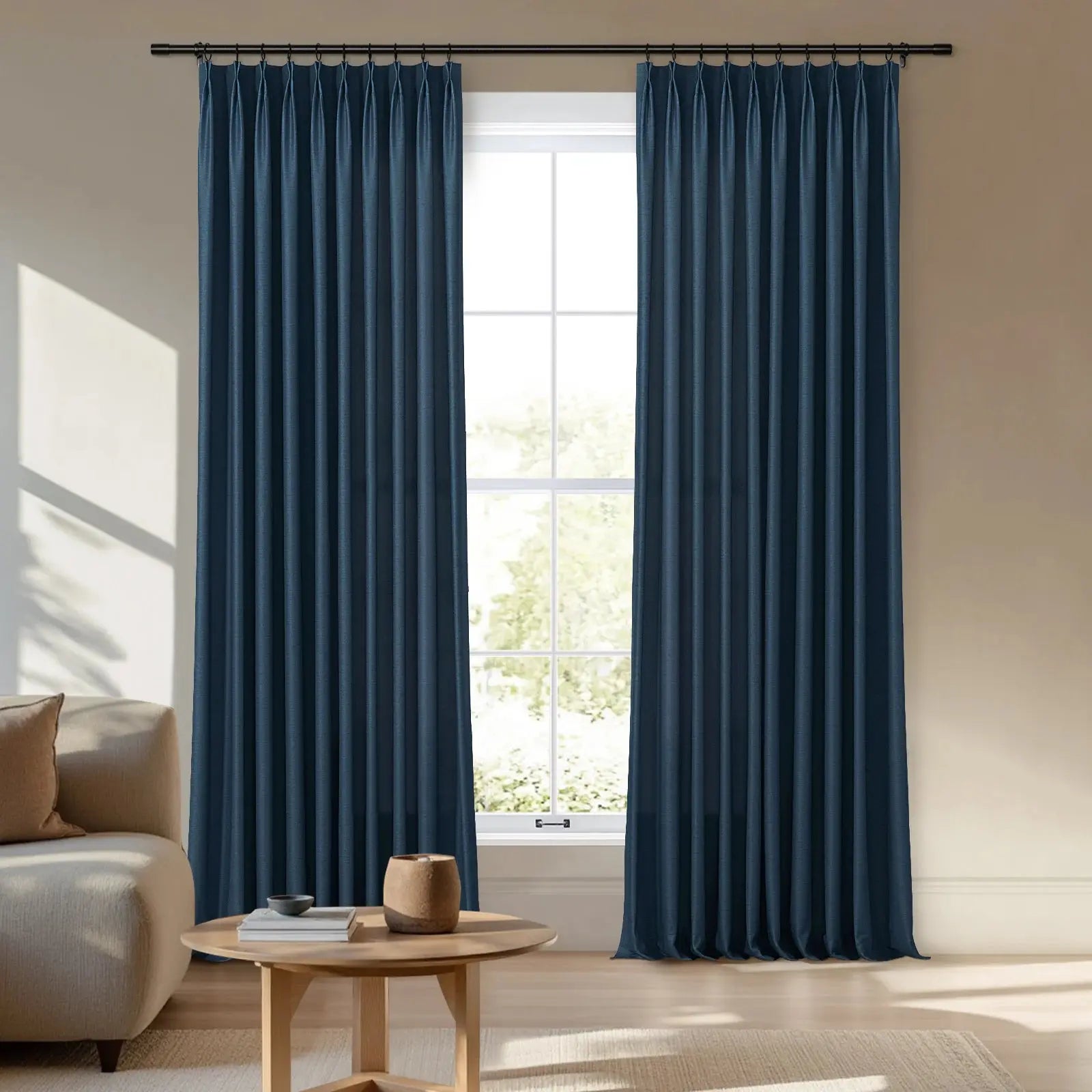 Photographic image of a contemporary living room featuring a pair of deep indigo pleated curtains on a black rod framing a window looking out onto lush greenery with a beige sofa and wooden coffee table in natural light
