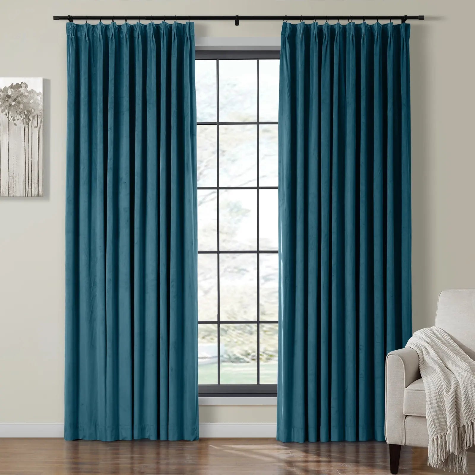 Photograph of two rich teal velvet curtain panels on a black rod framing a window with black grid panes, accompanied by a grayscale tree artwork on the left and a beige armchair with a white woven throw on the right