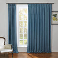 A cozy room interior featuring full-length teal blue linen-textured curtains on a black rod framing a white grid-paned window revealing sunlit green trees, alongside a cream upholstered armchair with a carved dark wood frame and a framed botanical lemon print on the right wall