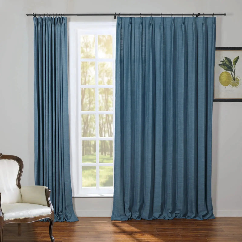 A cozy room interior featuring full-length teal blue linen-textured curtains on a black rod framing a white grid-paned window revealing sunlit green trees, alongside a cream upholstered armchair with a carved dark wood frame and a framed botanical lemon print on the right wall