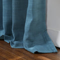 Close-up of a teal blue linen-textured curtain with gathered folds resting on a brown wooden floor beside a white wall trim