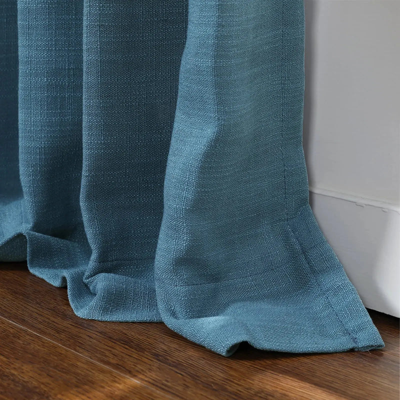 Close-up of a teal blue linen-textured curtain with gathered folds resting on a brown wooden floor beside a white wall trim