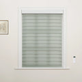 Photograph of a window in an interior room featuring gray-green banded zebra roller Blinds, a white remote control mounted on the right wall, and a small framed botanical artwork on the left wall