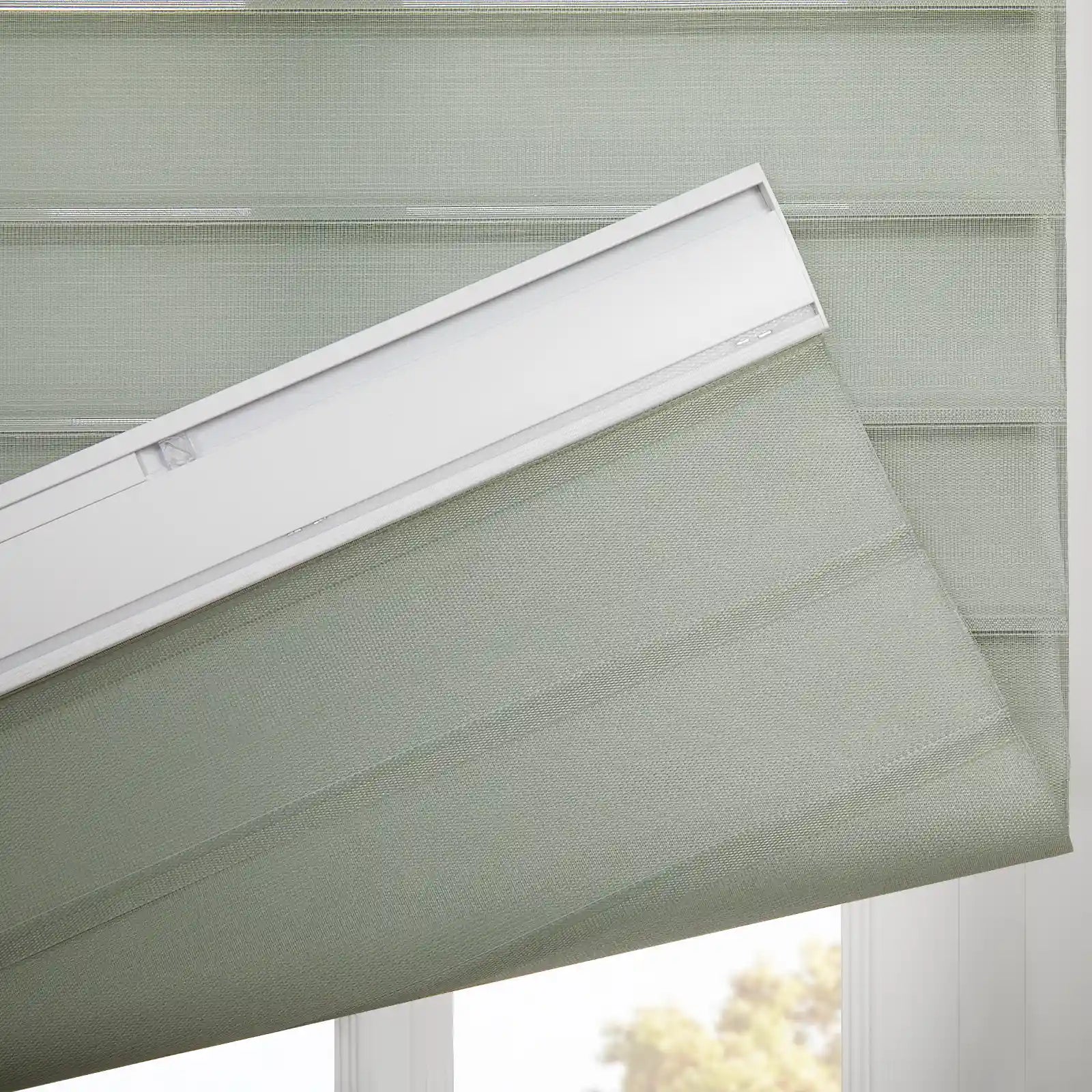 A light sage green dual-layer window Blind featuring alternating sheer and opaque horizontal stripes with a woven texture, partially unfurled from its white headrail at a window revealing blurred outdoor foliage