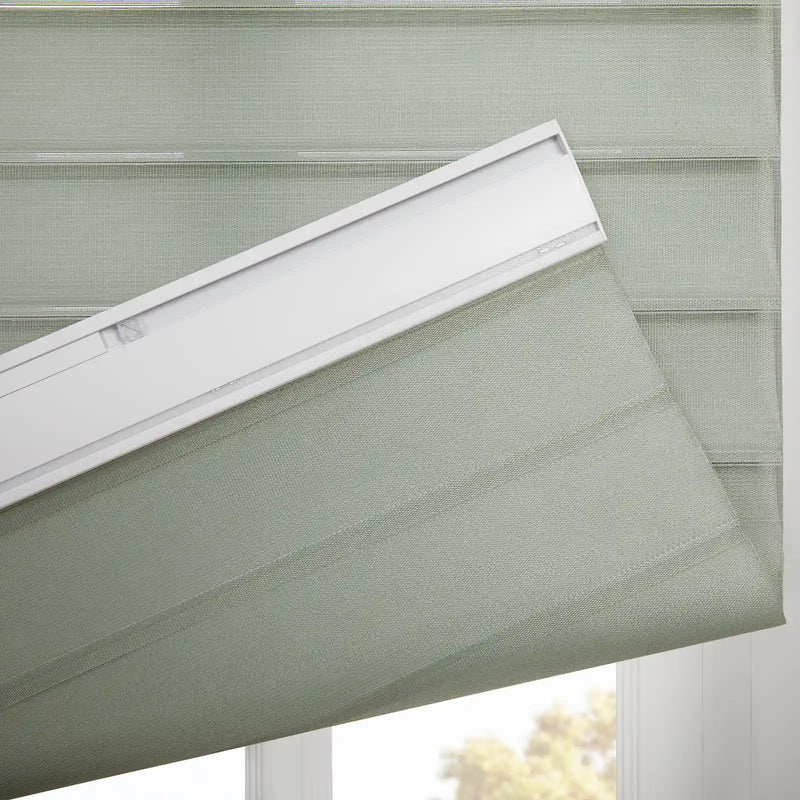 A light sage green dual-layer window Blind featuring alternating sheer and opaque horizontal stripes with a woven texture, partially unfurled from its white headrail at a window revealing blurred outdoor foliage