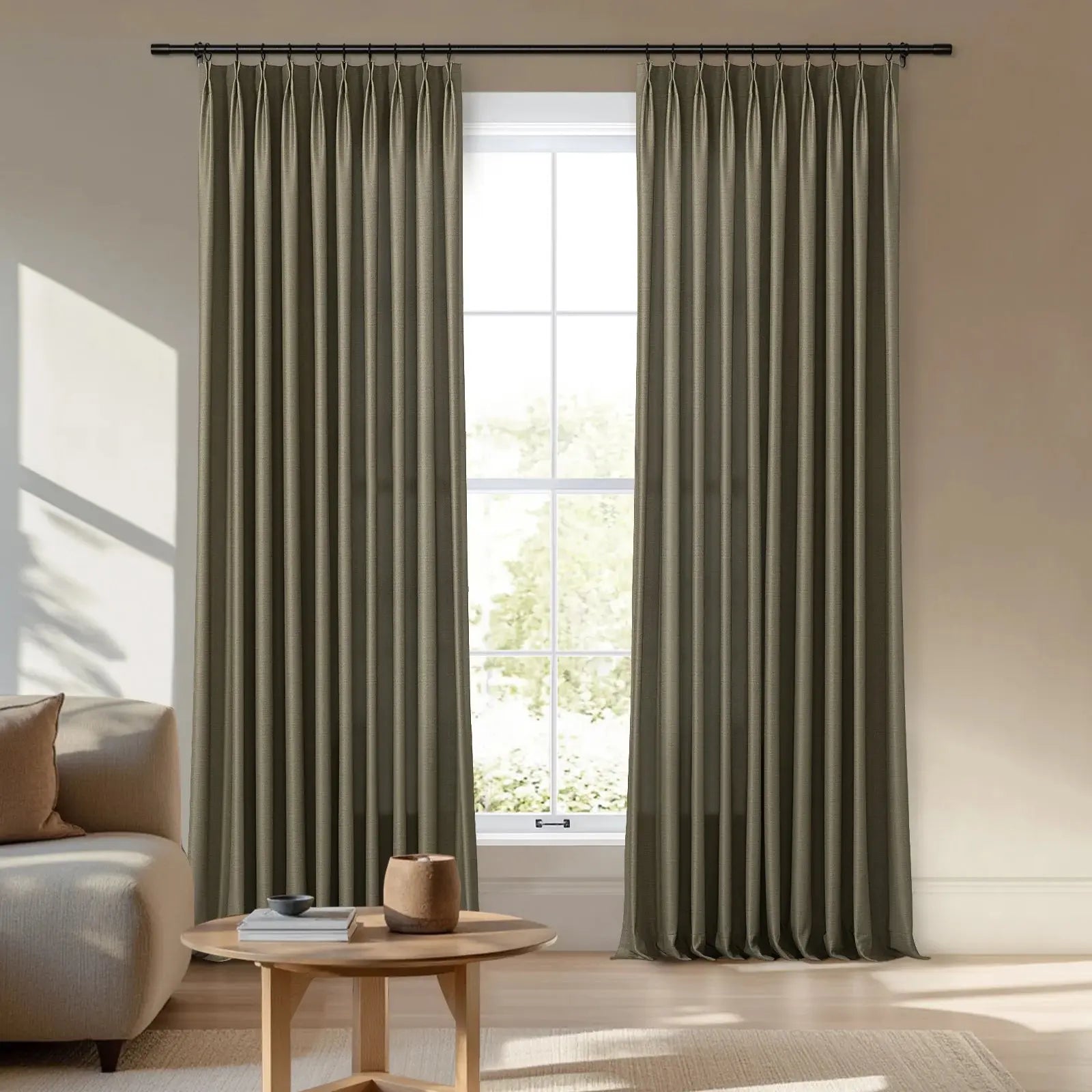 Photograph of a contemporary living room with olive green pinch-pleat drapes on a black rod framing a window with bright outdoor foliage, showcasing a beige sofa with a brown pillow and a light wooden coffee table with books and a rustic pot, all under sunlit geometric wall shadows