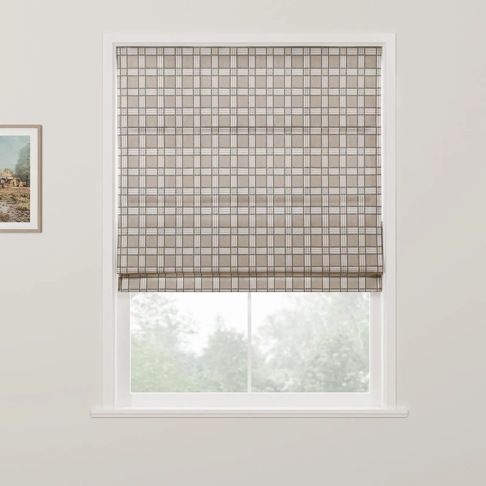 Photograph of a window with a beige and white plaid Roman blind partially covering a blurred view of green trees outside, accompanied by a small framed rustic landscape painting on the white wall to the left
