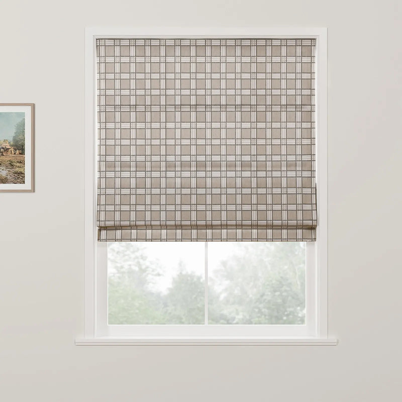 Photograph of a window with a beige and white plaid Roman blind partially covering a blurred view of green trees outside, accompanied by a small framed rustic landscape painting on the white wall to the left