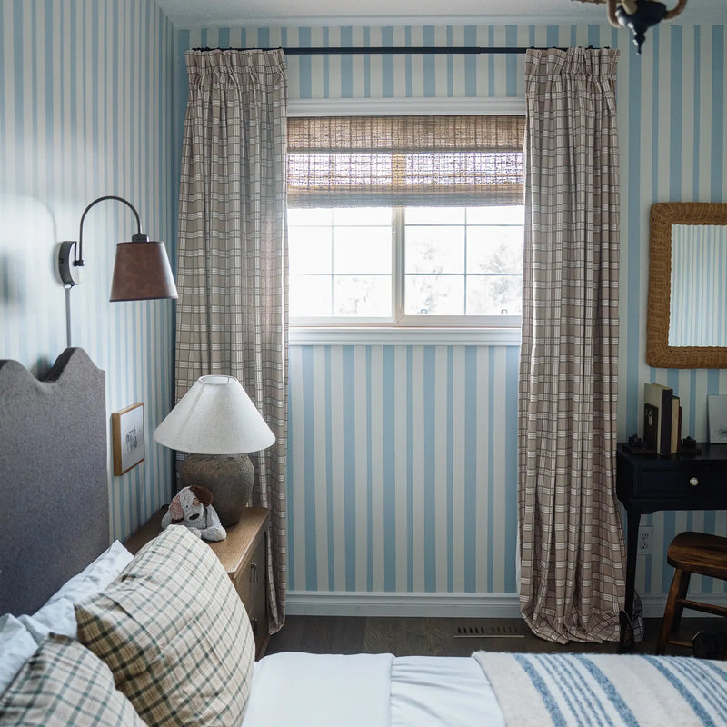 A cozy bedroom featuring blue and white striped wallpaper, plaid curtains and bedding, a woven wood blind, and a plush dog toy on the nightstand