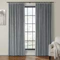 Grey velvet pinch-pleat curtains frame a black grid window in a bright modern living room featuring an abstract grey tree artwork and a cream textured throw blanket on a beige armchair