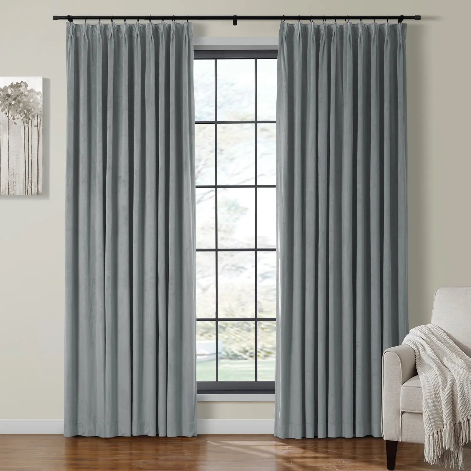 Grey velvet pinch-pleat curtains frame a black grid window in a bright modern living room featuring an abstract grey tree artwork and a cream textured throw blanket on a beige armchair