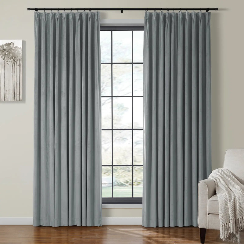 Grey velvet pinch-pleat curtains frame a black grid window in a bright modern living room featuring an abstract grey tree artwork and a cream textured throw blanket on a beige armchair