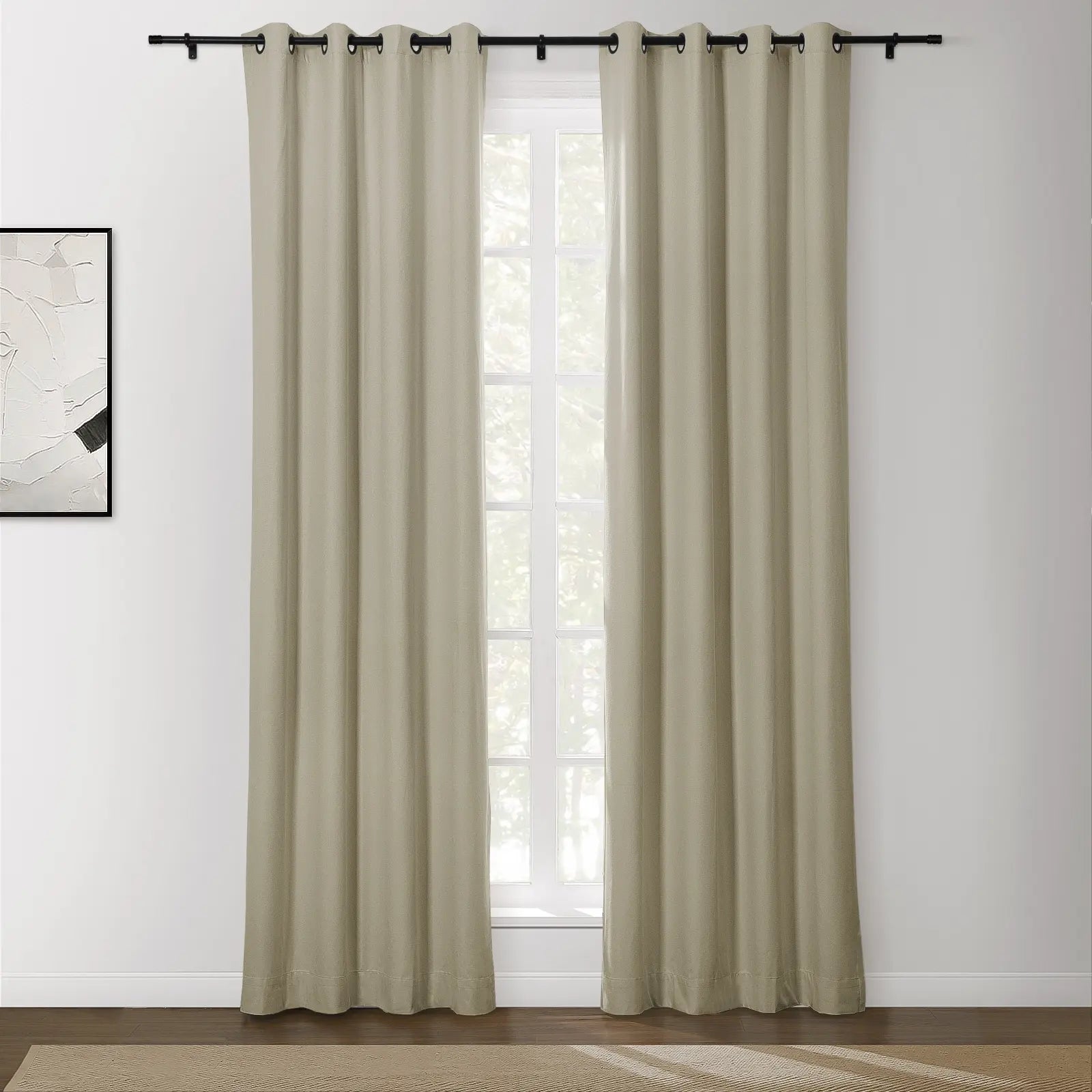 Beige grommet top curtains on a black rod in a bright room with an abstract white and black framed artwork and a view of outdoor greenery through the window above a natural fiber rug
