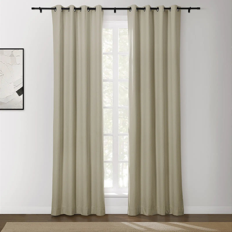 Beige grommet top curtains on a black rod in a bright room with an abstract white and black framed artwork and a view of outdoor greenery through the window above a natural fiber rug