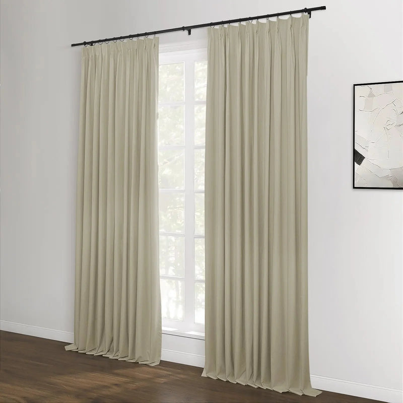 Beige pinch-pleat curtains on a black rod frame a large white-framed window with a lush tree view, set against a white wall with abstract black and white artwork and a dark wood floor