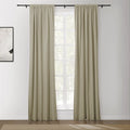 A pair of pleated beige curtains on a black rod over a multi-pane window with visible green foliage in a modern room featuring abstract black and white framed artwork and a textured tan rug on a wooden floor