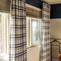 Interior view of a sunlit window framed by blue and beige plaid curtains, tan cellular shades, a dark blue accent wall, and a partial wooden valet stand