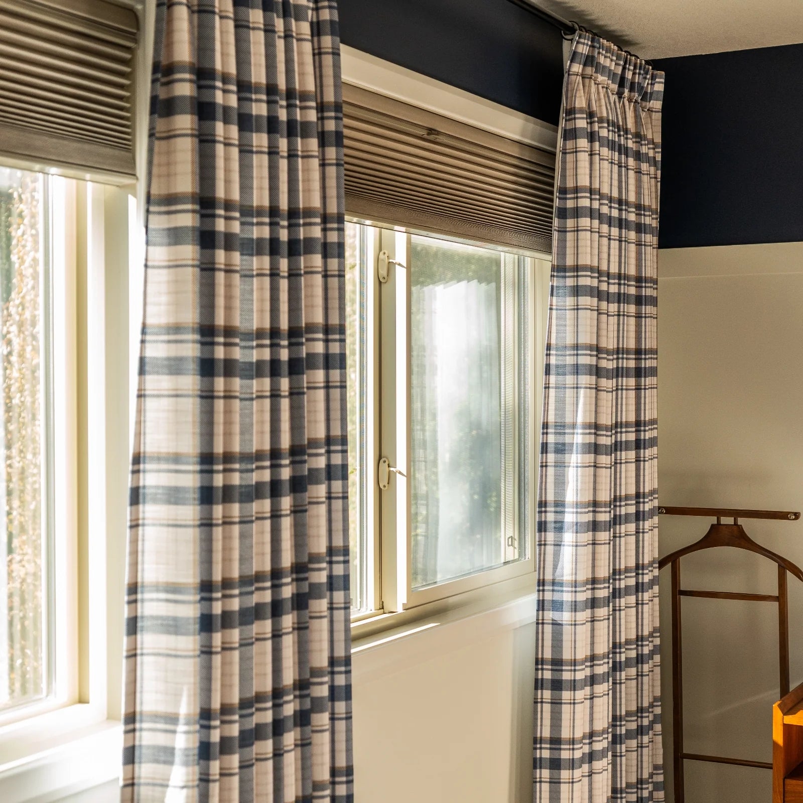 Interior view of a sunlit window framed by blue and beige plaid curtains, tan cellular shades, a dark blue accent wall, and a partial wooden valet stand
