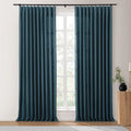 Photograph of deep teal blue pleated curtains on a black rod in a bright room featuring a textured linen-like fabric, with a fluted wooden side table holding a black vase with greenery and a white candle on a gold holder