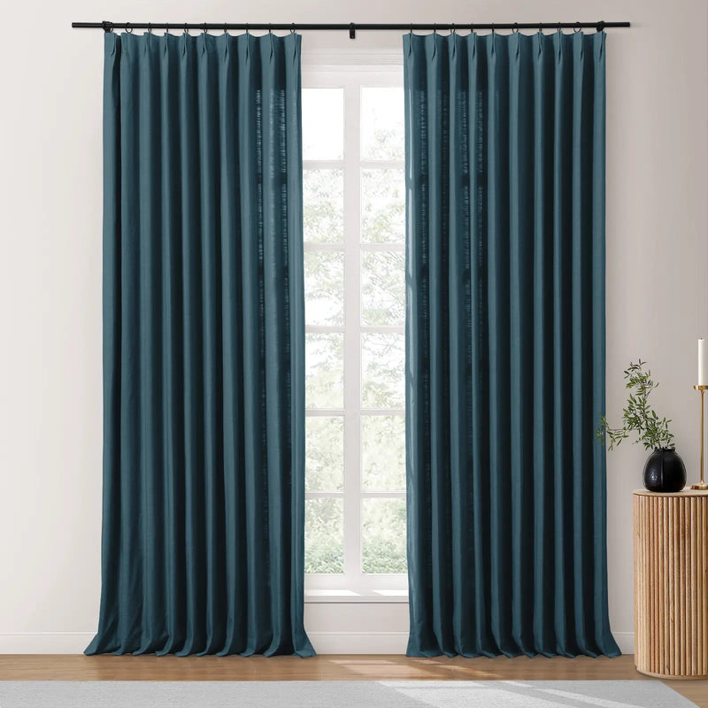 Photograph of deep teal blue pleated curtains on a black rod in a bright room featuring a textured linen-like fabric, with a fluted wooden side table holding a black vase with greenery and a white candle on a gold holder