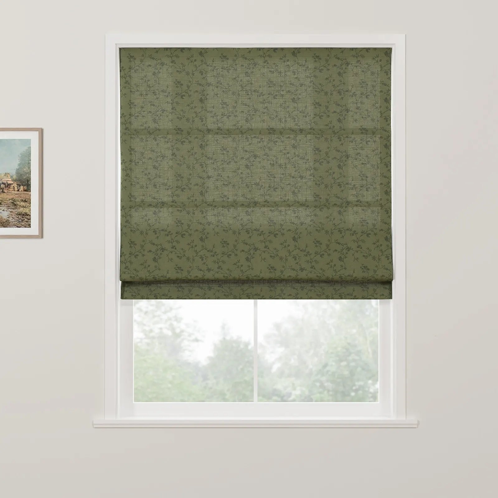 A Roman blind in olive green with a subtle grey floral and grid pattern covers the top of a white-framed window, with blurred green trees visible outside and a framed landscape painting on the adjacent wall