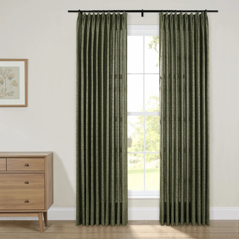 Pleated dark green patterned curtains on a black rod adorn a bright window in a contemporary room featuring a light wood mid-century modern dresser with brass pulls and a framed botanical print artwork