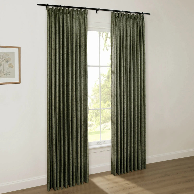Photograph of a bright room with a window framed by dark olive green pleated curtains featuring a subtle pattern, a black rod, a botanical print on the wall, and light wood floors