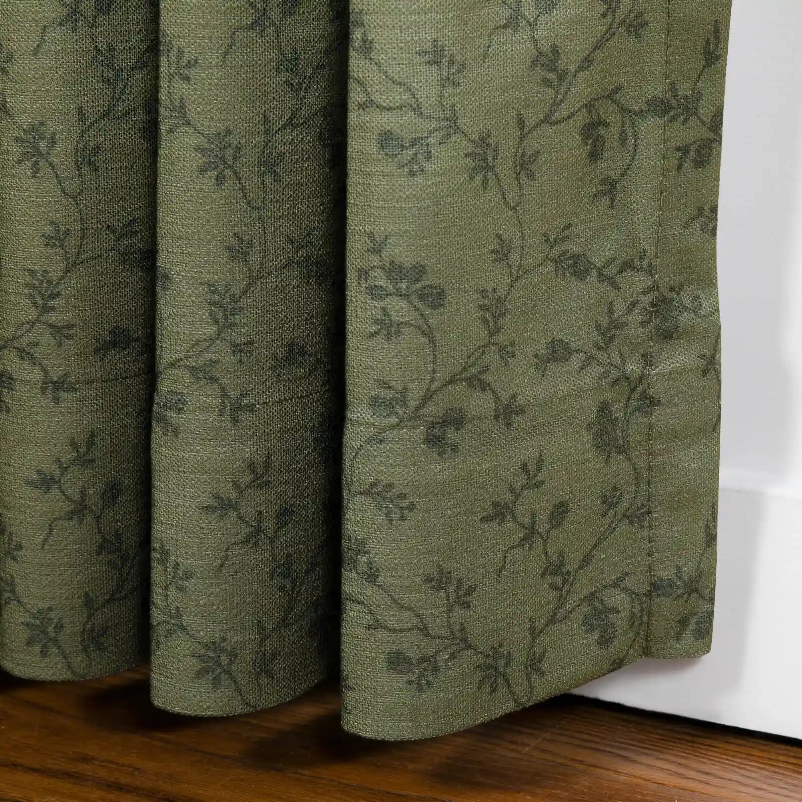 A muted olive green textile curtain with a subtle dark grey botanical vine pattern showing visible woven texture and soft folds rests on a rich brown wooden floor against a white baseboard