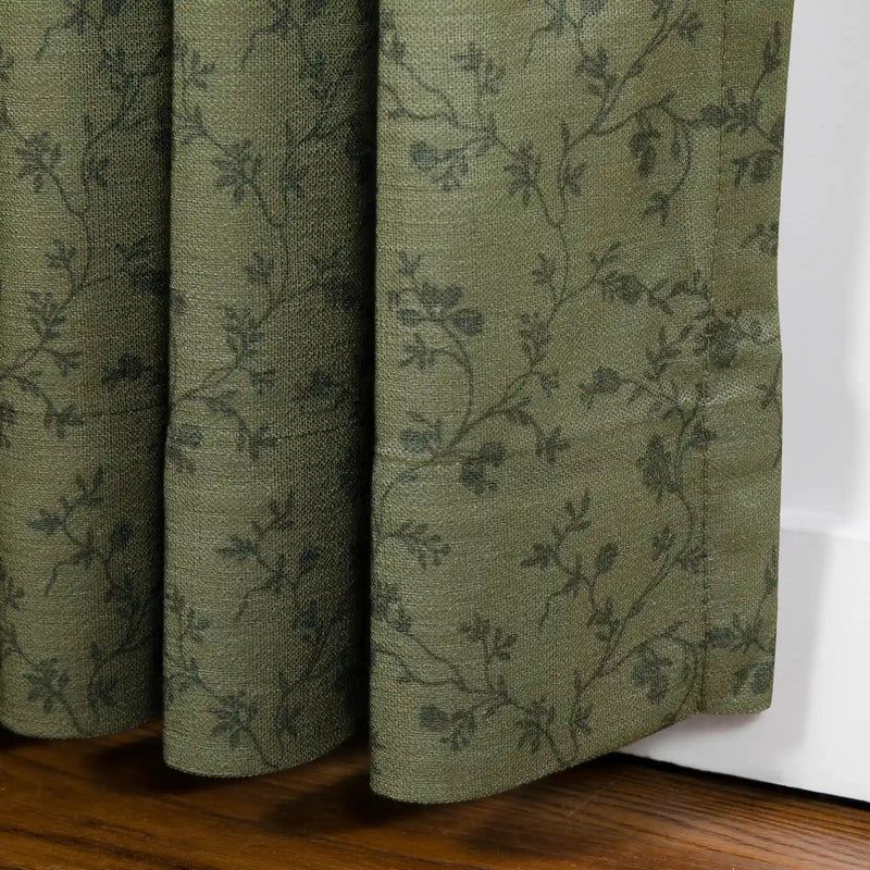 A muted olive green textile curtain with a subtle dark grey botanical vine pattern showing visible woven texture and soft folds rests on a rich brown wooden floor against a white baseboard
