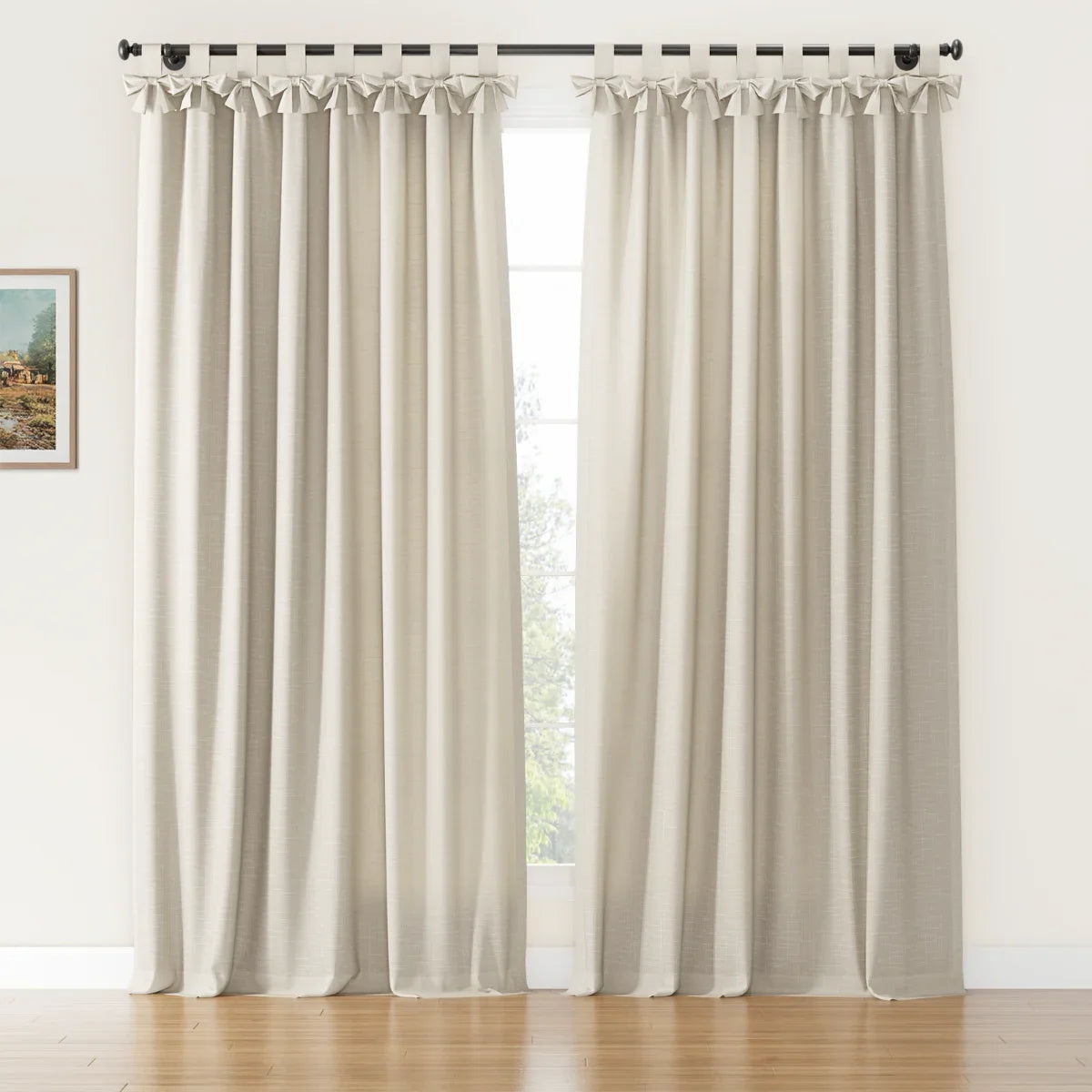 A pair of textured light beige tie-top curtains with decorative bow details hangs on a black rod covering a window with outdoor greenery in a room with a wooden floor and a small framed landscape painting on the left wall