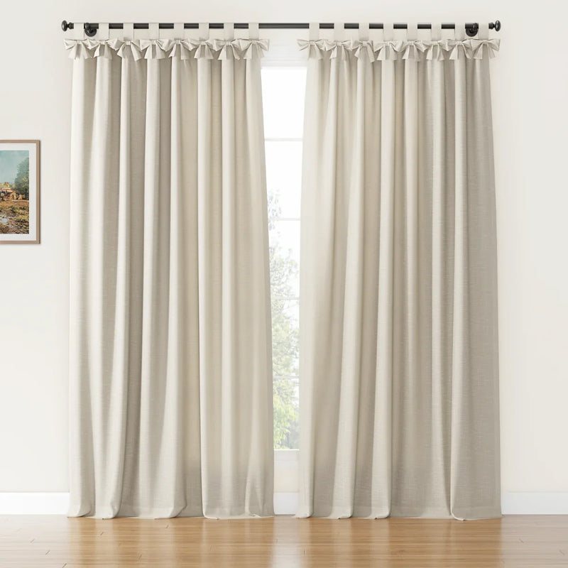A pair of textured light beige tie-top curtains with decorative bow details hangs on a black rod covering a window with outdoor greenery in a room with a wooden floor and a small framed landscape painting on the left wall