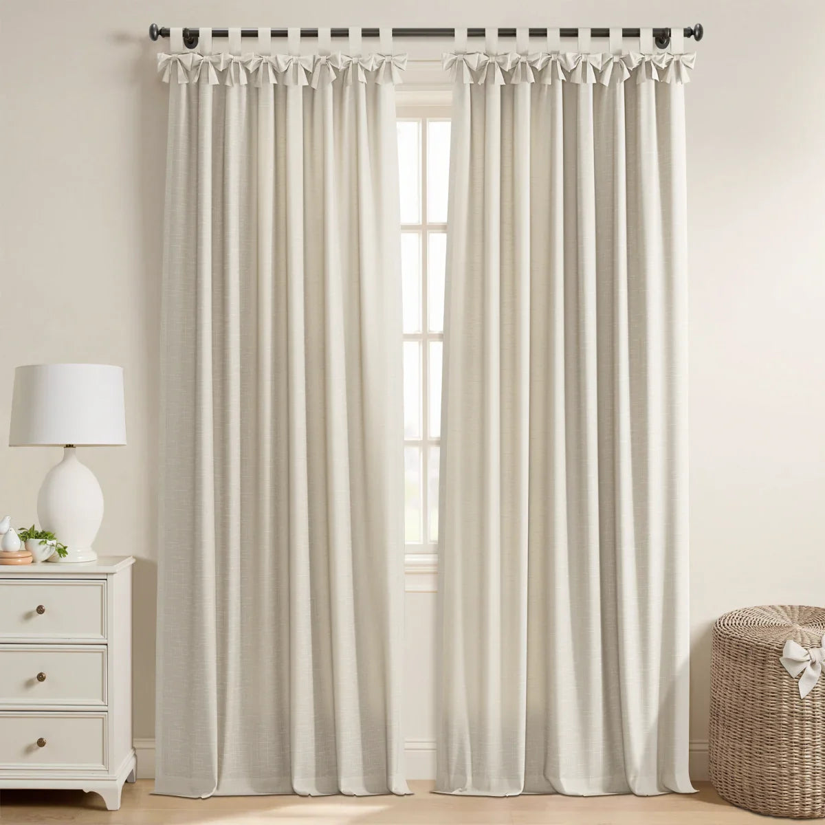 Photograph of a cozy room featuring full-length neutral beige linen window curtains with elegant tab tops and bow accents, a white three-drawer nightstand with a matching ceramic lamp, and a woven wicker basket with a white bow