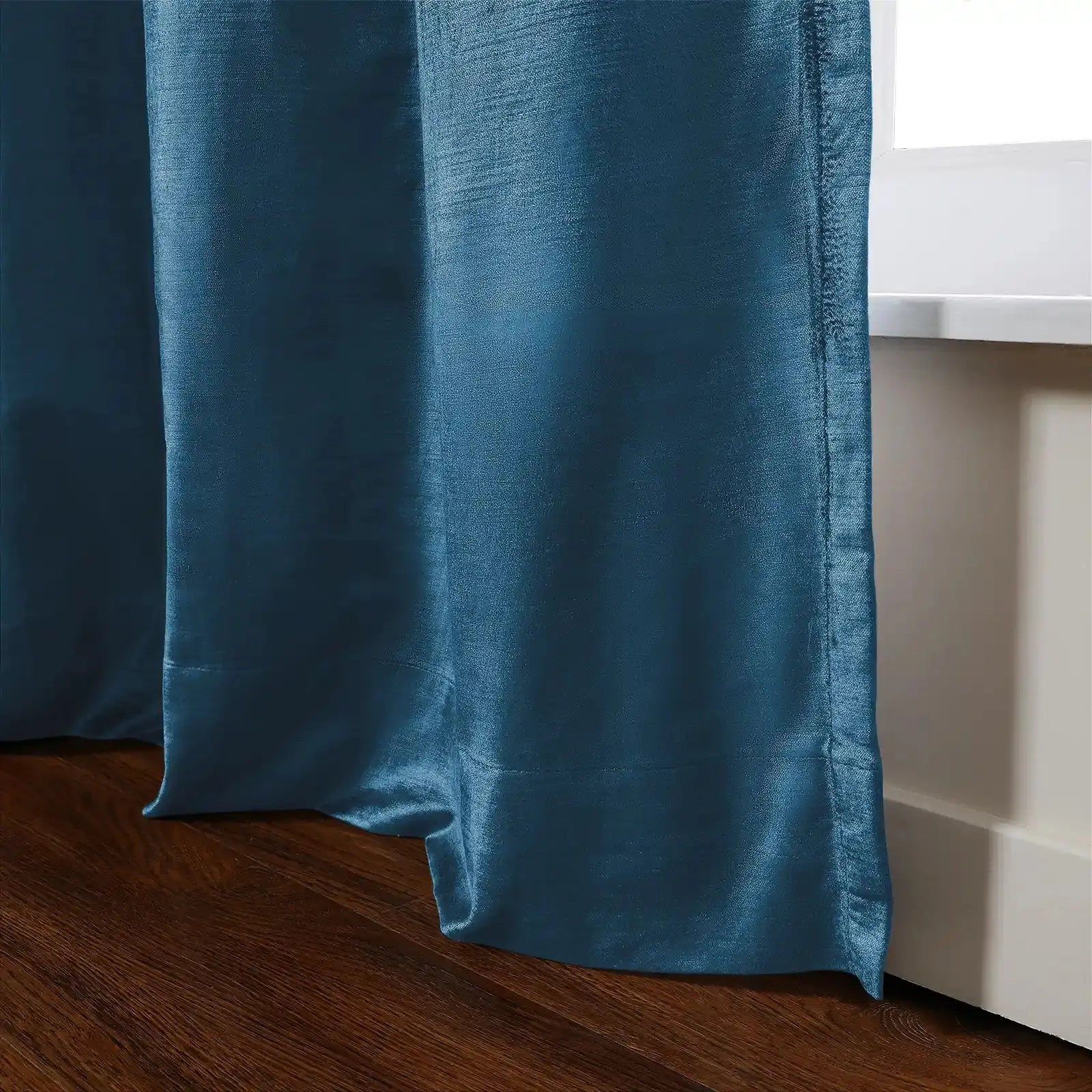A rich teal blue curtain with a subtle sheen and visible weave elegantly drapes onto a dark wooden floor next to a white window sill revealing soft folds
