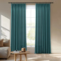 A pair of vibrant teal pinch-pleat curtains frame a sunlit window in a contemporary living room featuring a light beige sofa and a round wooden coffee table