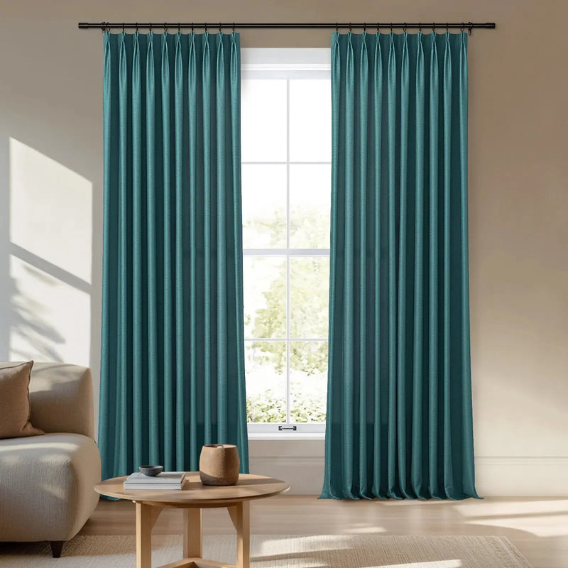 A pair of vibrant teal pinch-pleat curtains frame a sunlit window in a contemporary living room featuring a light beige sofa and a round wooden coffee table