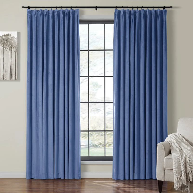 A pair of rich blue velvet pleated curtains frame a grid-patterned window in a home interior, flanked by an abstract gray tree art piece and a beige armchair with a textured throw