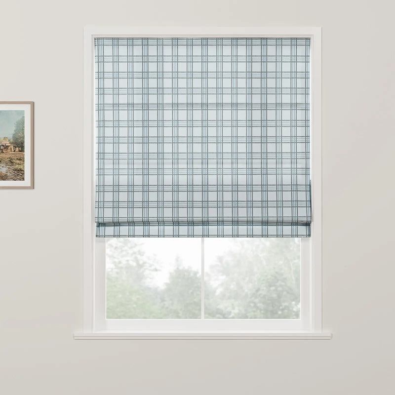 Photograph of a white-framed window featuring a light blue and dark blue plaid Roman blind neatly folded at the bottom, with a partially visible framed landscape print on the left wall and blurred green trees outside