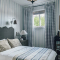 A cozy bedroom interior with blue and white striped wallpaper, plaid curtains and pillows, a wavy gray upholstered headboard, and a soft blue striped blanket
