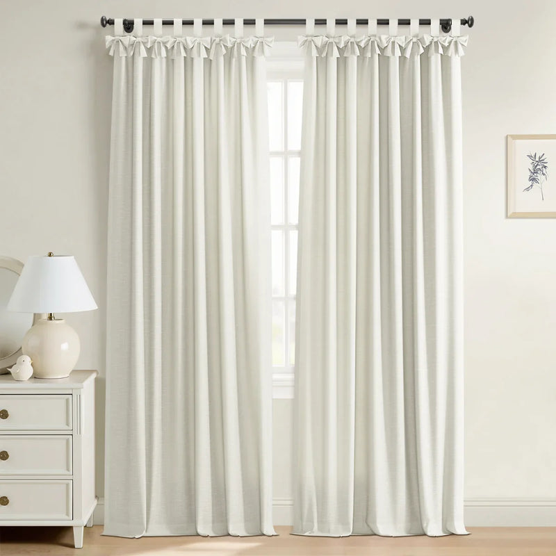 White textured tab-top curtains with decorative bows on a window in a bright room featuring a white three-drawer dresser with brass knobs, a ceramic lamp, and a framed botanical print