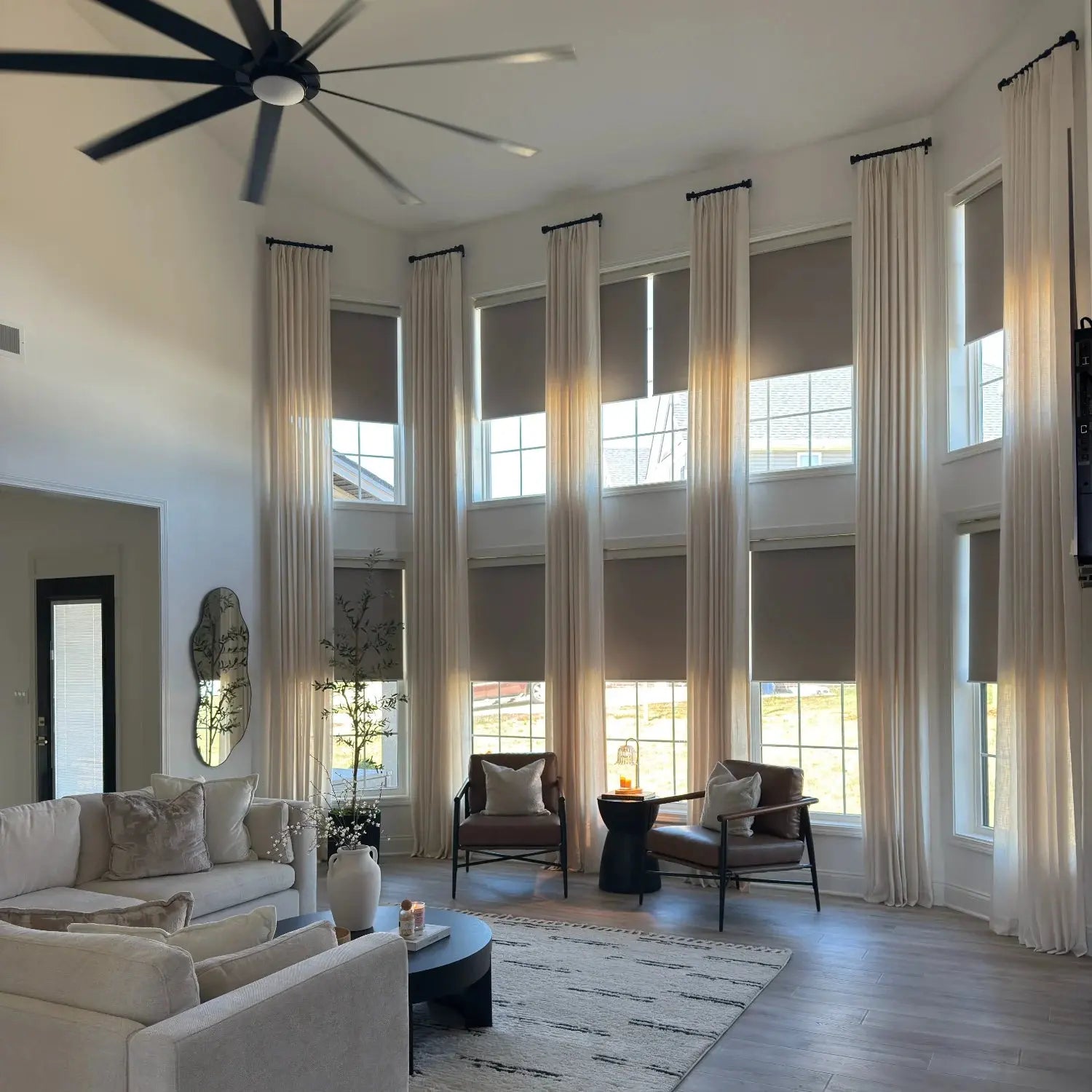 Photograph of a modern spacious living room featuring a large black multi-blade ceiling fan, tall windows with cream floor-to-ceiling drapes and dark roller shades, a cream sectional sofa, dark leather armchairs, and a distinctive wavy gold mirror