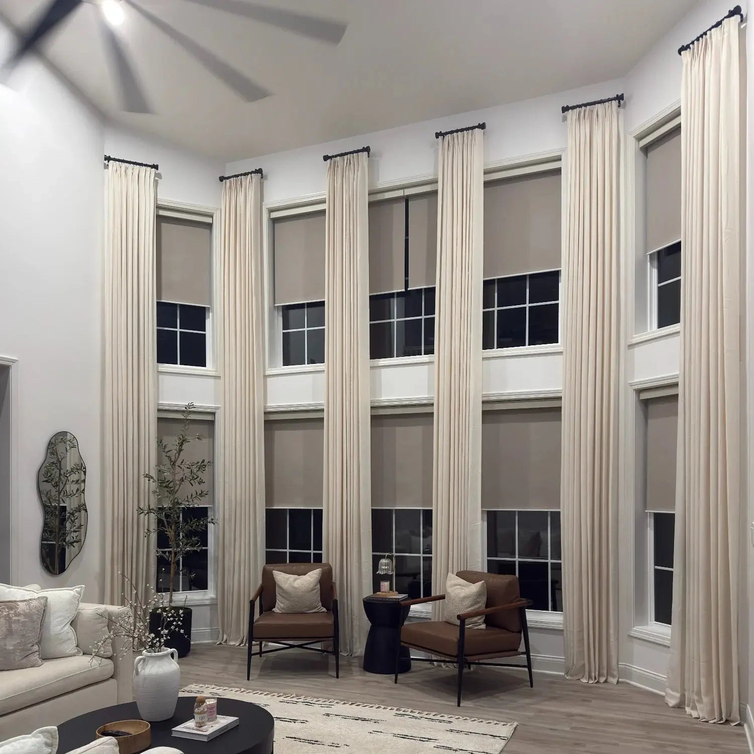 Photograph of a modern living room interior with multiple tall windows adorned with beige drapes and blackout roller shades, complemented by two brown leather armchairs, an irregularly shaped mirror, and a ceiling fan