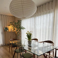 A brightly lit modern dining room interior featuring a large white Akari-style paper lantern, a glass-top dining table with a potted yellow daffodil plant, mid-century modern wooden chairs, and a fluted light wood cabinet displaying abstract art against sheer white curtains