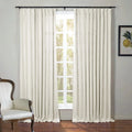 An interior scene featuring full-length cream pleated curtains on a black rod, a sunlit window revealing outdoor trees, a classic white upholstered armchair with dark wood trim, and a framed lemon botanical print
