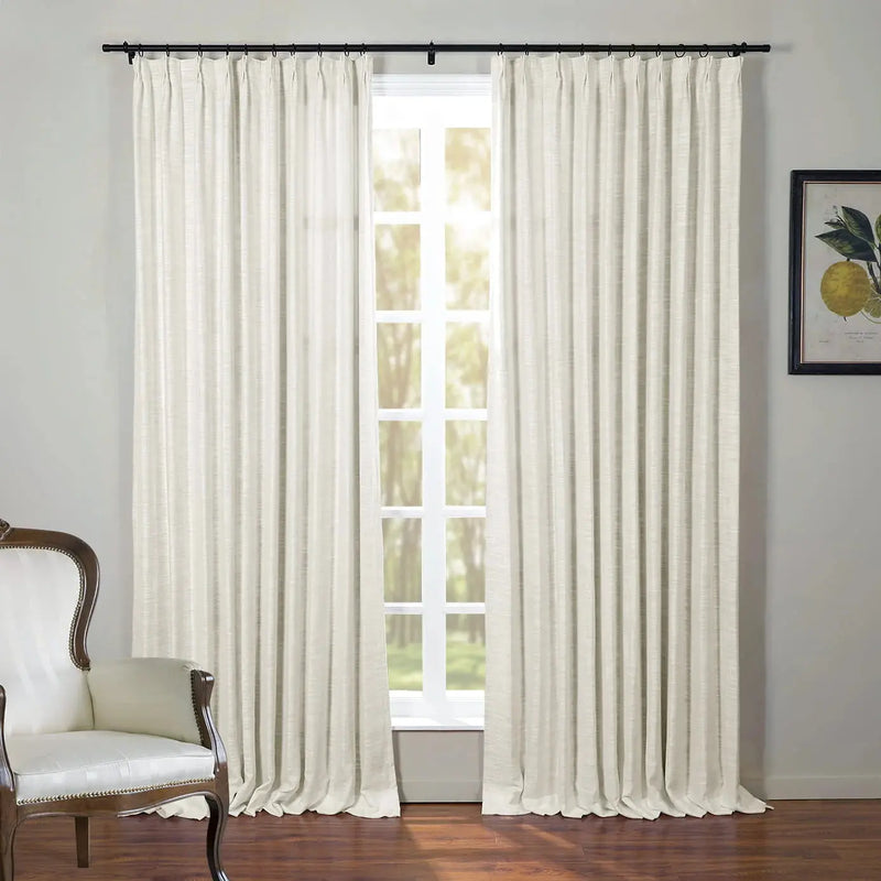 An interior scene featuring full-length cream pleated curtains on a black rod, a sunlit window revealing outdoor trees, a classic white upholstered armchair with dark wood trim, and a framed lemon botanical print