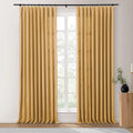 Mustard yellow linen-look curtains hanging from a black rod over a white-gridded window, with green foliage visible outside and a ribbed wooden side table featuring a black vase and white candle