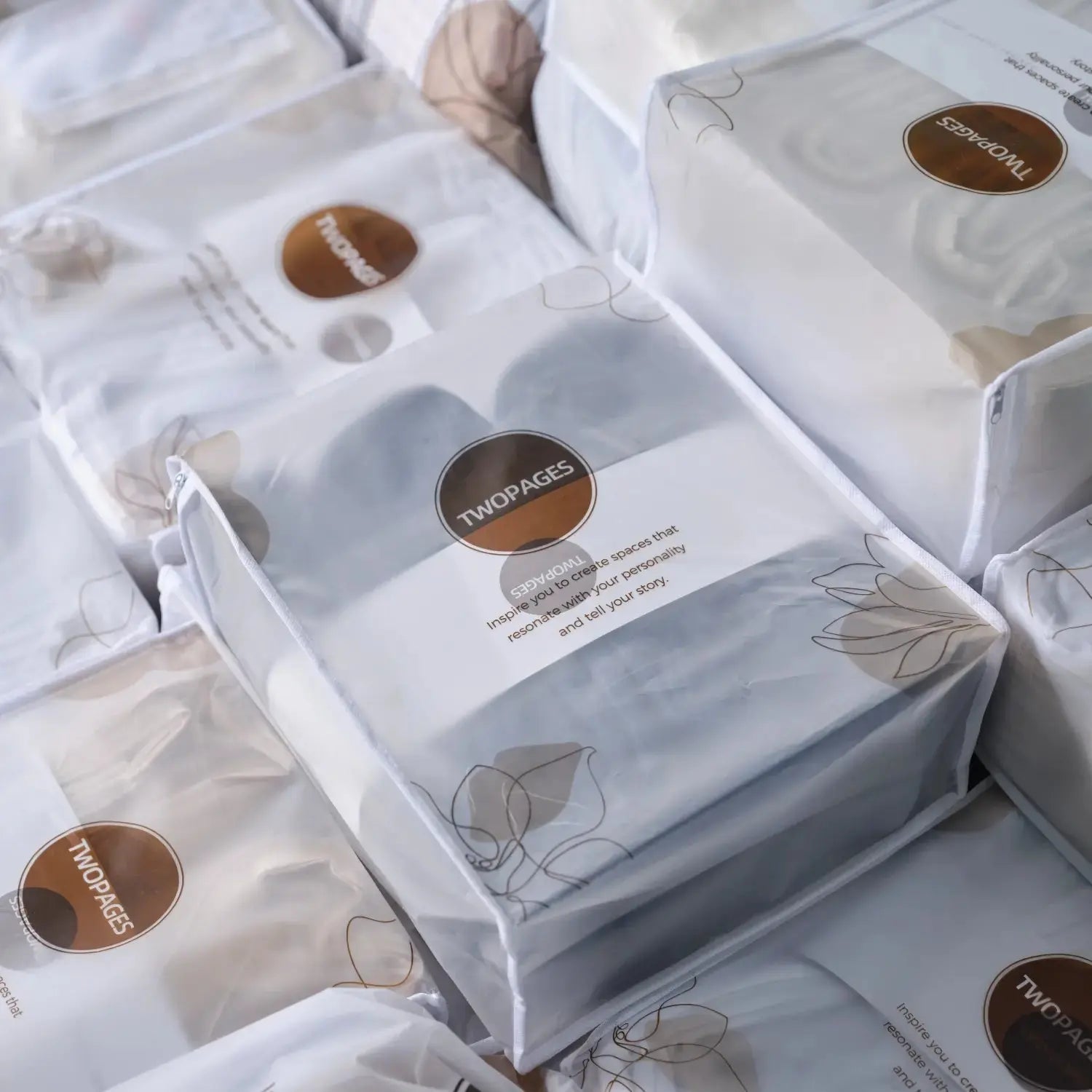 A close-up of multiple white translucent zippered TWOPAGES packages holding rolled textiles, each package featuring a brown circular logo, subtle abstract floral line art, and the slogan Inspire you to create spaces