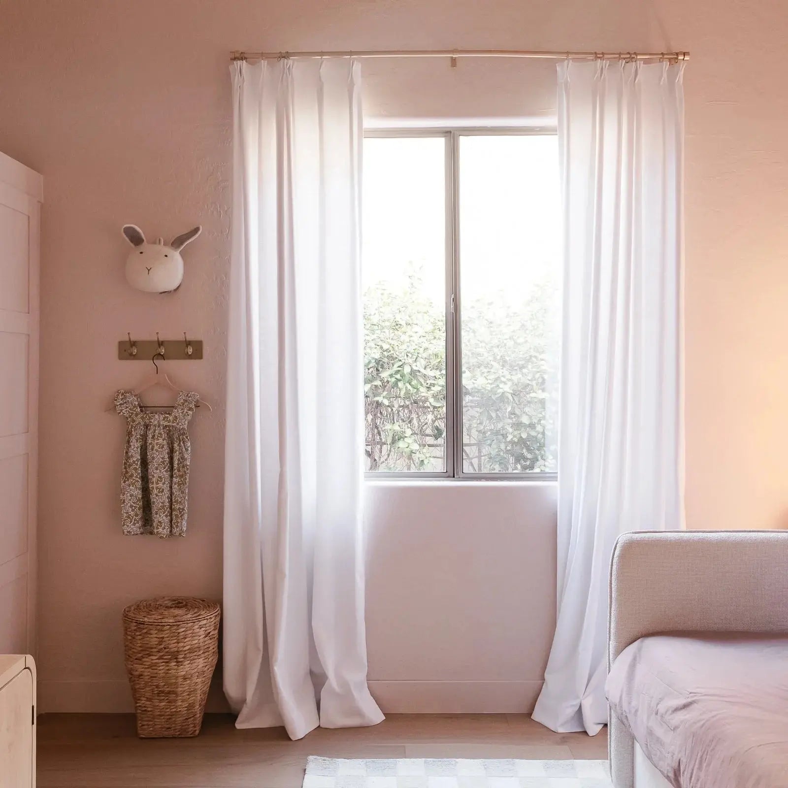 A tranquil children's bedroom with soft pink walls, bright white curtains framing a window overlooking green foliage, adorned with a white bunny head wall decor and a small floral dress on a hook