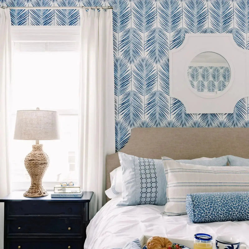 A bright coastal bedroom interior featuring blue palm leaf wallpaper a unique shaped white mirror a beige headboard with blue and white patterned pillows including a leopard print bolster and a woven lamp on a dark nightstand