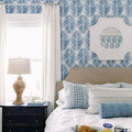 A bright coastal bedroom interior featuring blue palm leaf wallpaper a unique shaped white mirror a beige headboard with blue and white patterned pillows including a leopard print bolster and a woven lamp on a dark nightstand